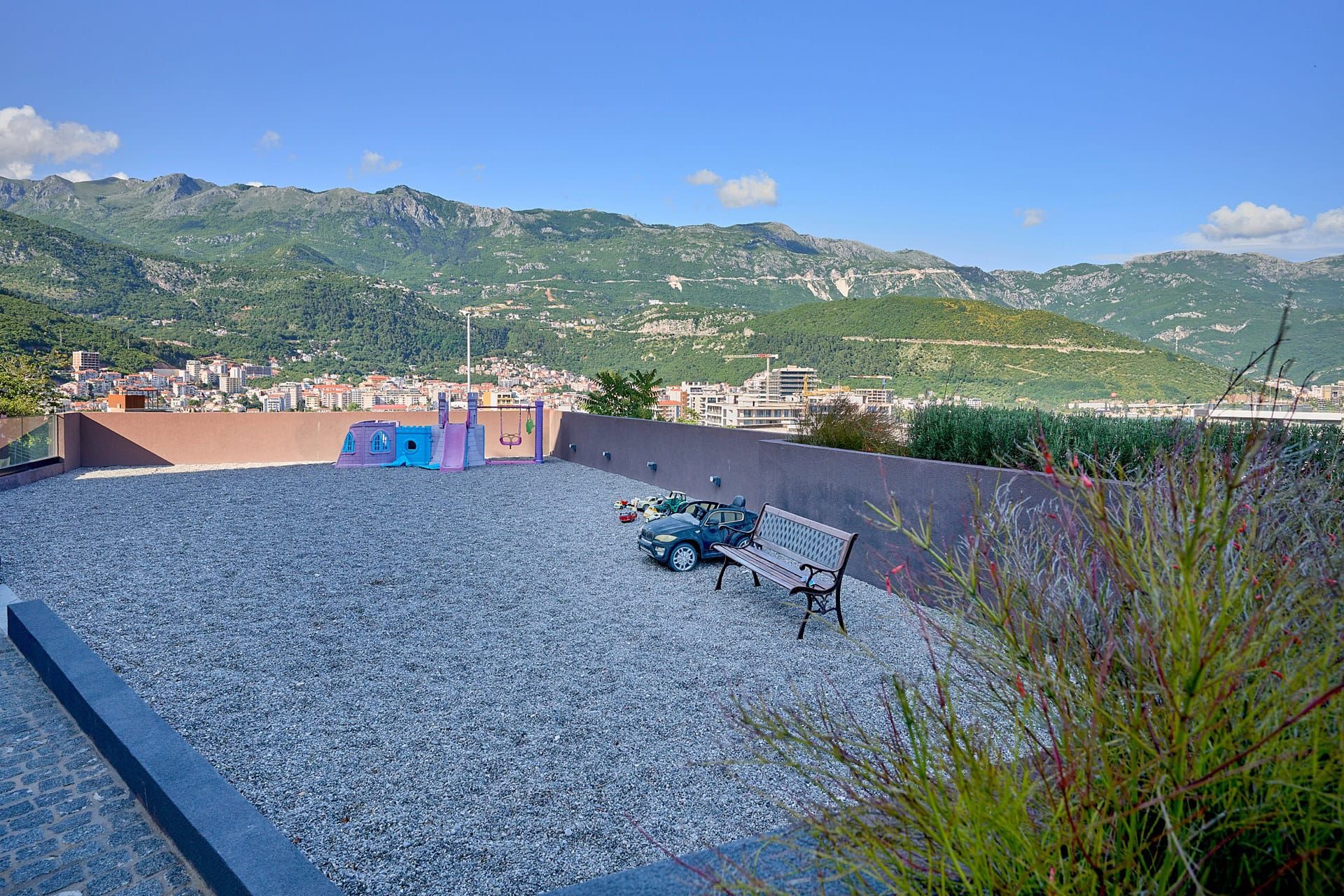 Terrace with a playground and mountain views in Budva, Montenegro: a bench, toys, and a city panorama.