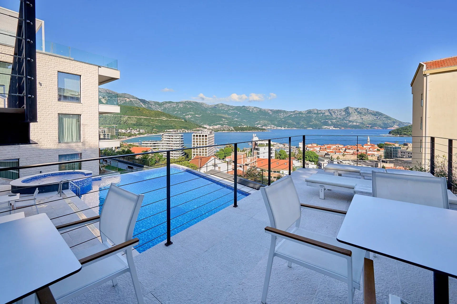 Rooftop pool with sea and mountain view, white furniture, jacuzzi, and glass railing.