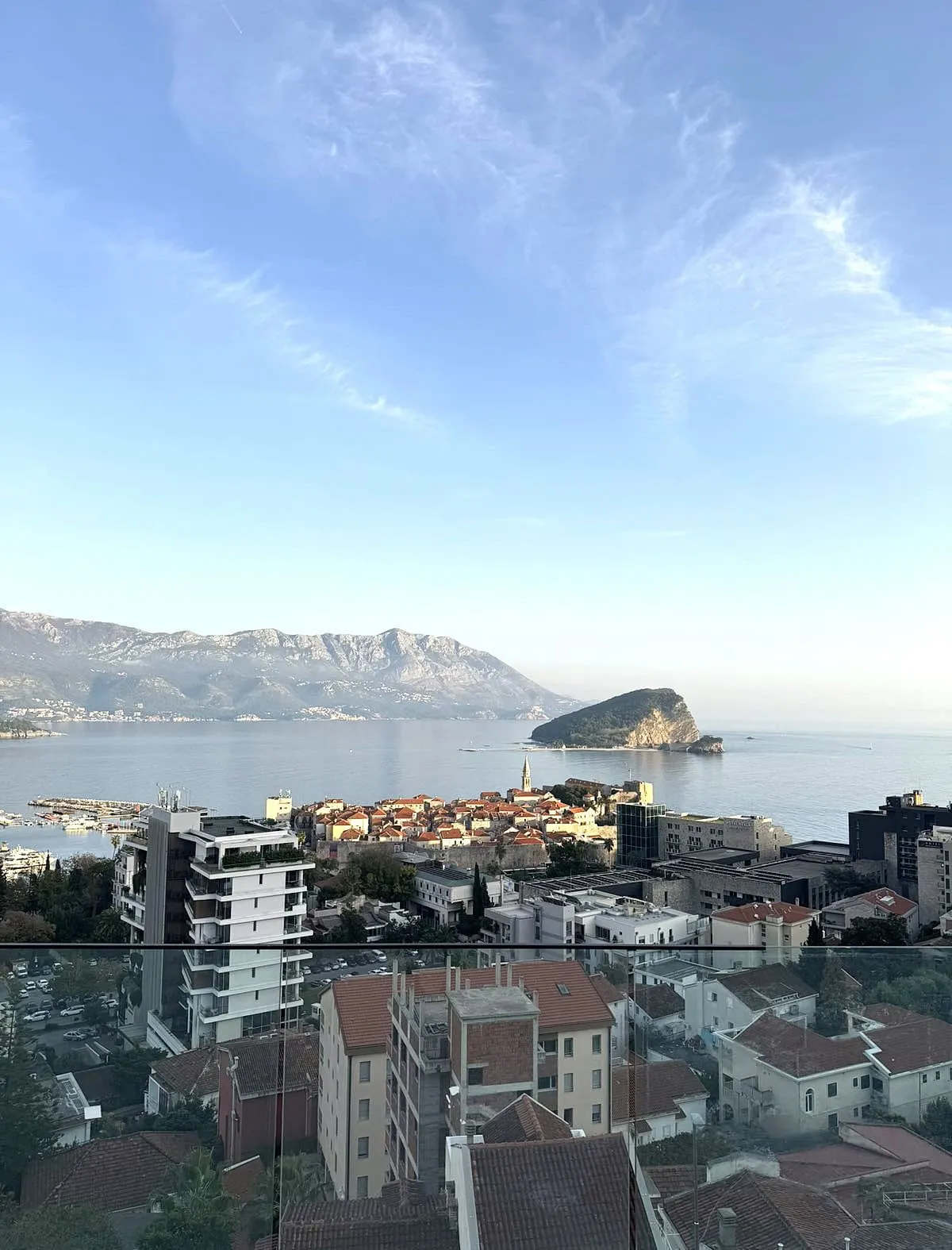 Panoramic view of a coastal city, sea, and mountains from a glass balcony.