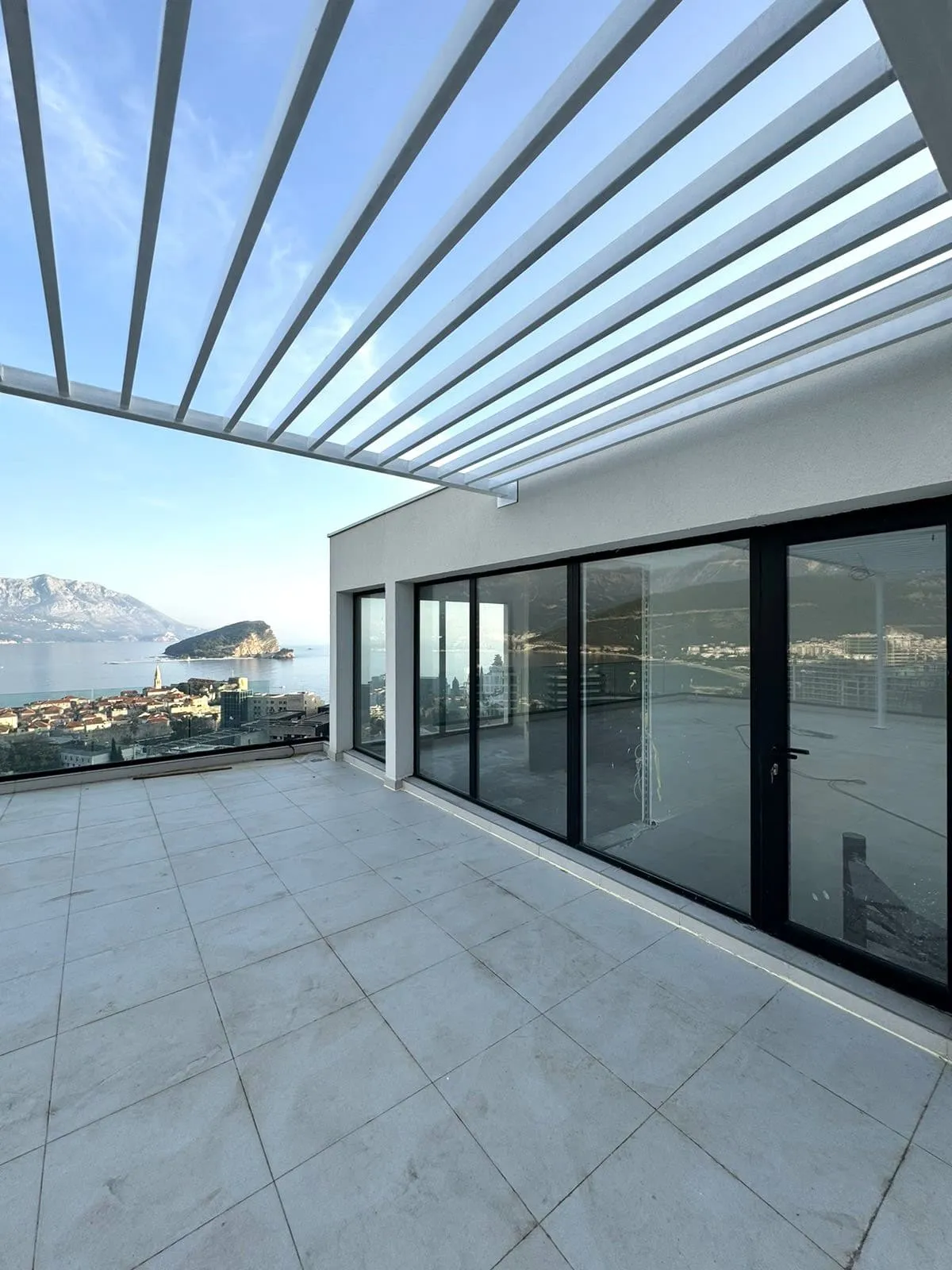 Wide tiled balcony with white pergola and glass doors. Panoramic view of the sea, city, and mountains.