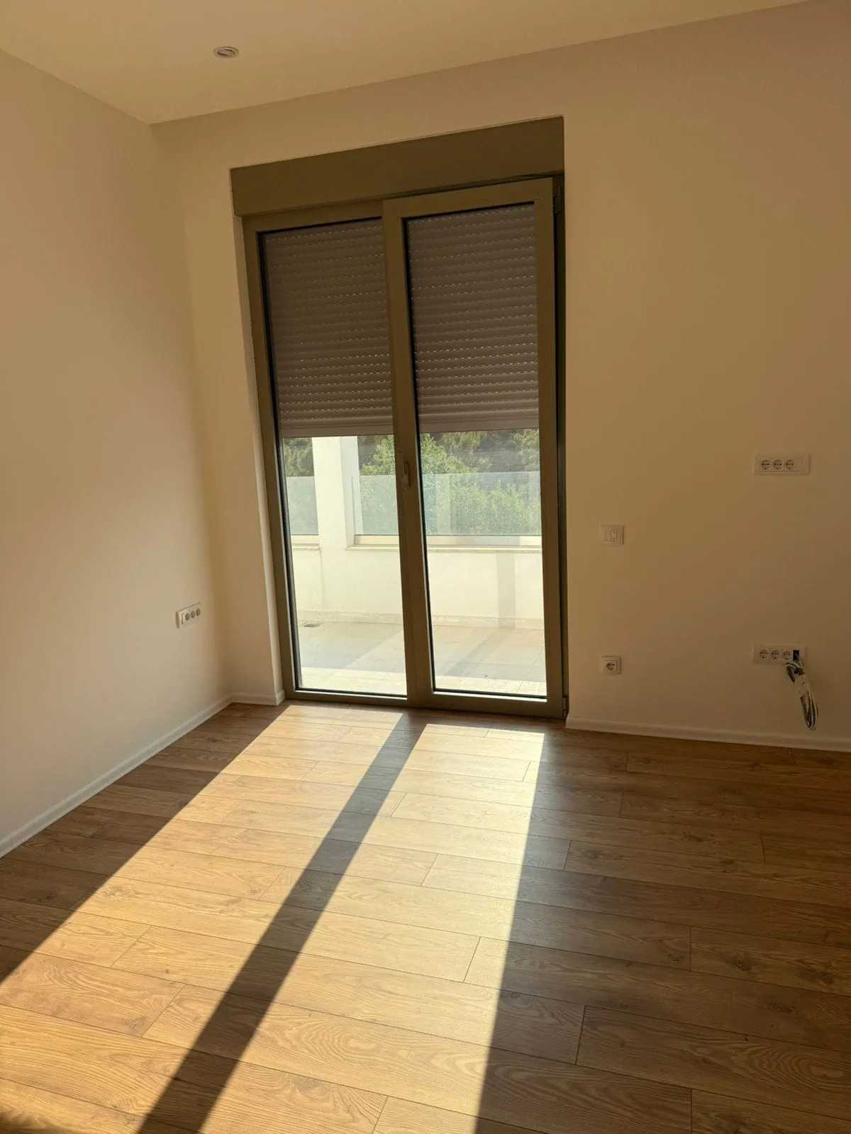 Empty room with wood laminate flooring and a glass door leading to a balcony with blinds.