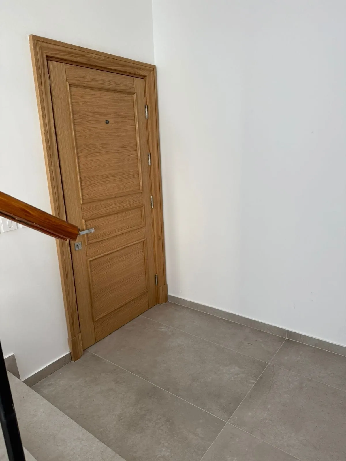 Wooden door with handle and hinges against white wall, gray tiled floor, and part of a wooden stair handrail visible on the left.
