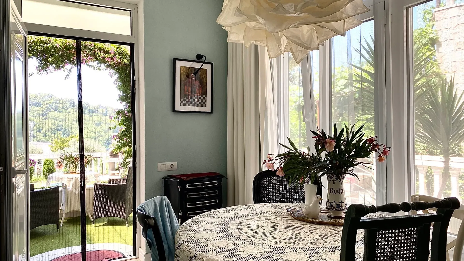 Dining area with a round lace-covered table, chairs, and a glass door opening to a garden balcony.