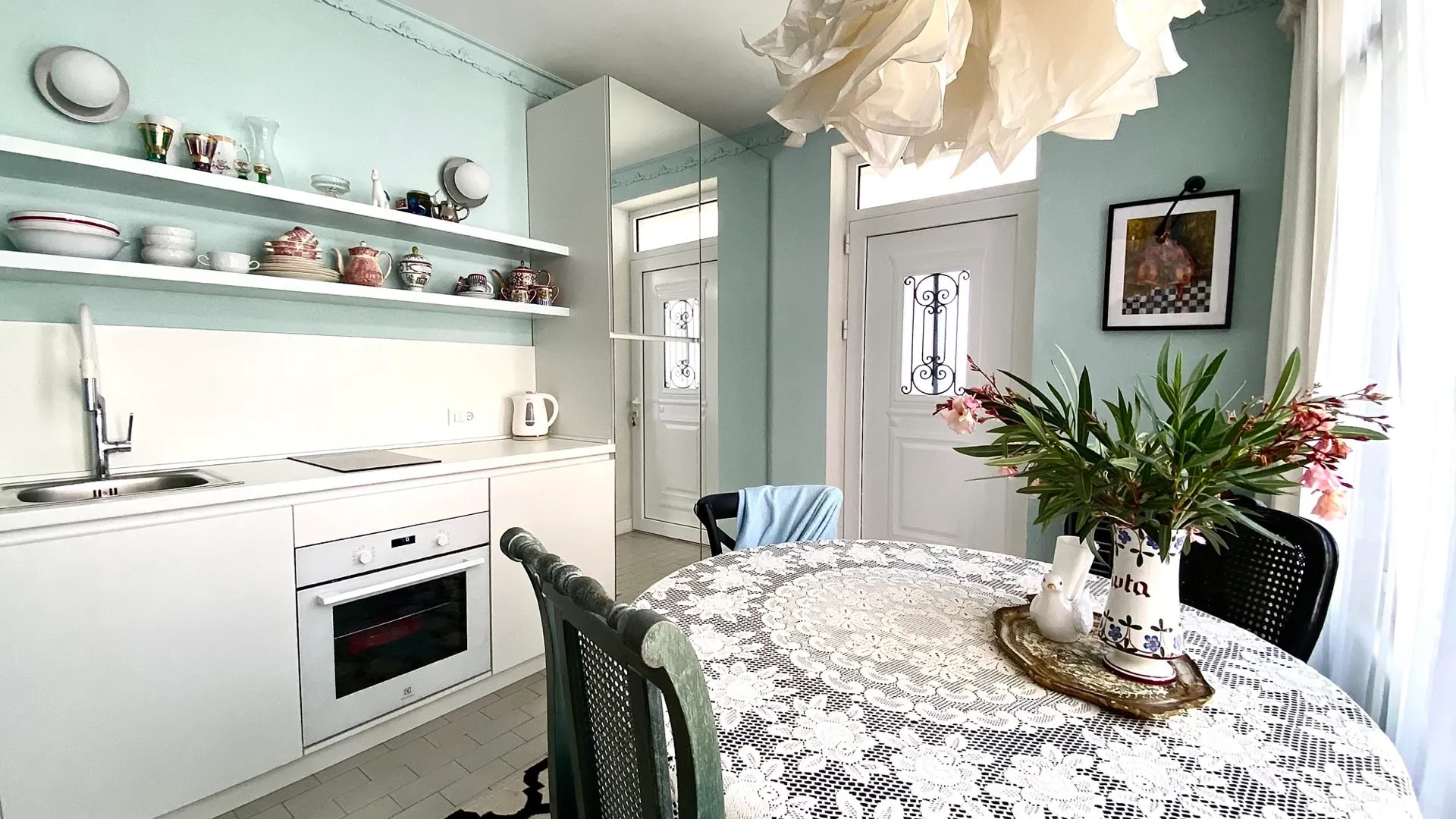 Kitchen with white cabinetry, sink, oven, and a round table covered in lace on light blue walls under a paper lamp.