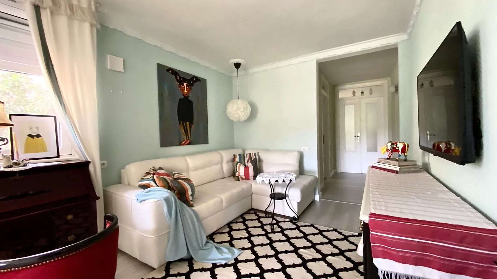 Living room with white sectional sofa, black and white rug, wall-mounted TV, and artwork on light green walls.
