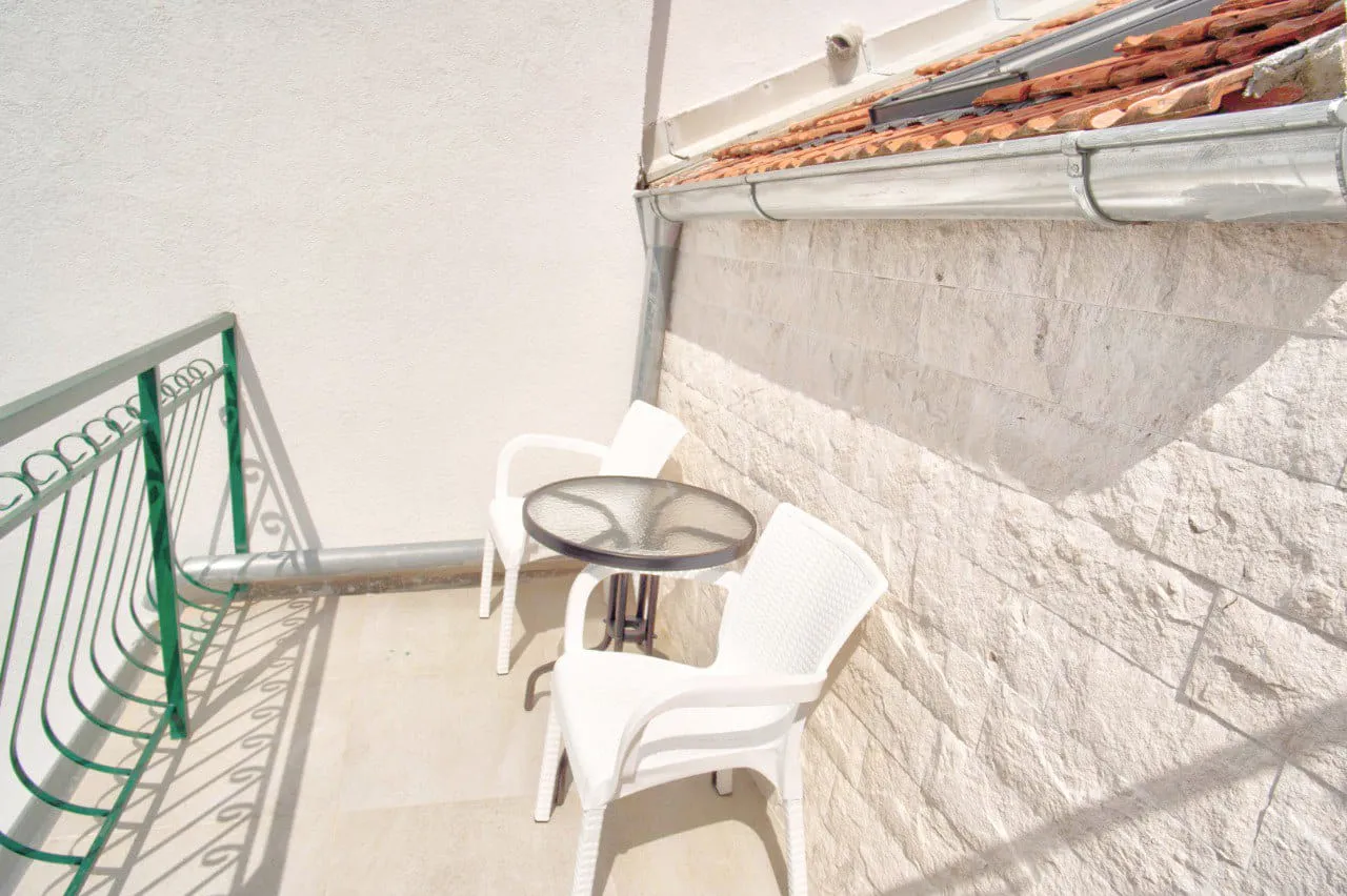 Small balcony with white plastic chairs, a round glass table, and a green metal railing against a stone wall.