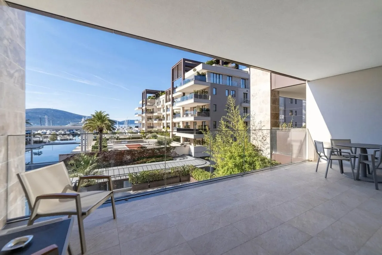 Wide balcony with glass railings, outdoor furniture, and a view of the marina and residential buildings.