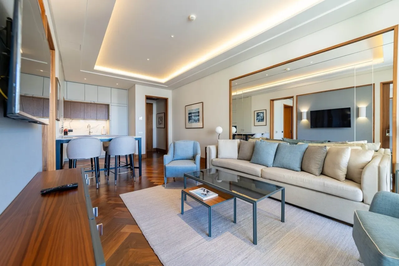 Modern living room with a beige sofa, large mirror, glass coffee table, and kitchen area in background.