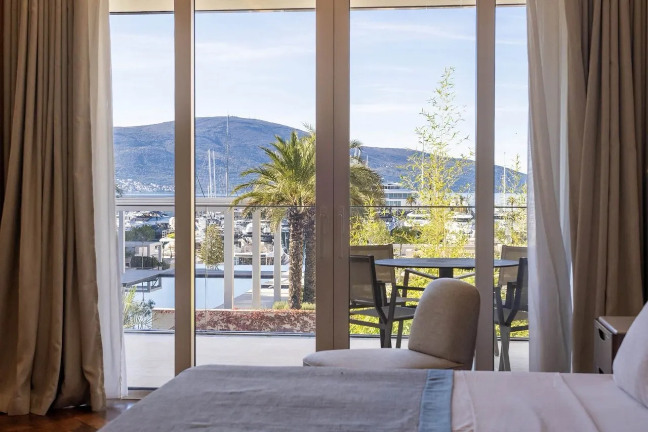 View from a bedroom showing a bed, glass doors leading to a balcony with table and chairs, overlooking a marina and mountains.