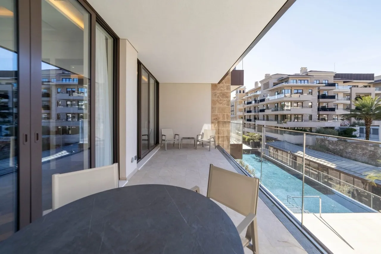 Modern balcony with dining set and glass railing overlooking a pool and residential buildings.