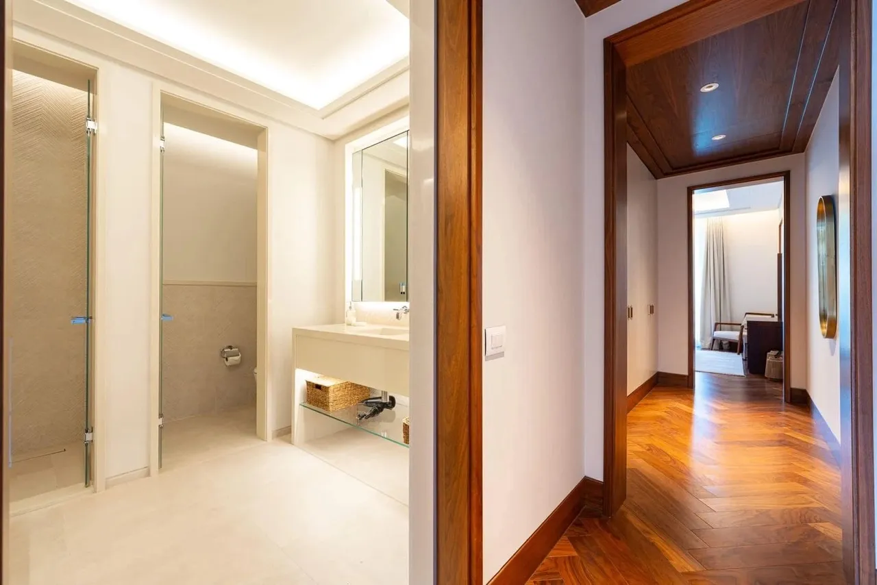 View of a bathroom with a glass shower, vanity sink, and mirror, connected to a wood-floored hallway.