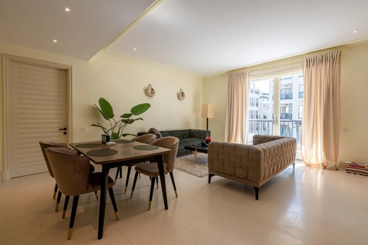 Living room with dining table, sofas, and balcony access. Bright interior with beige walls and light flooring.