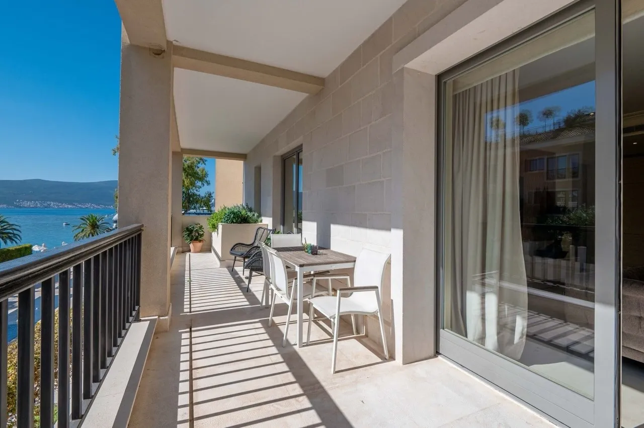 Balcony with a dining table and white chairs overlooking the sea and mountains through a metal railing.