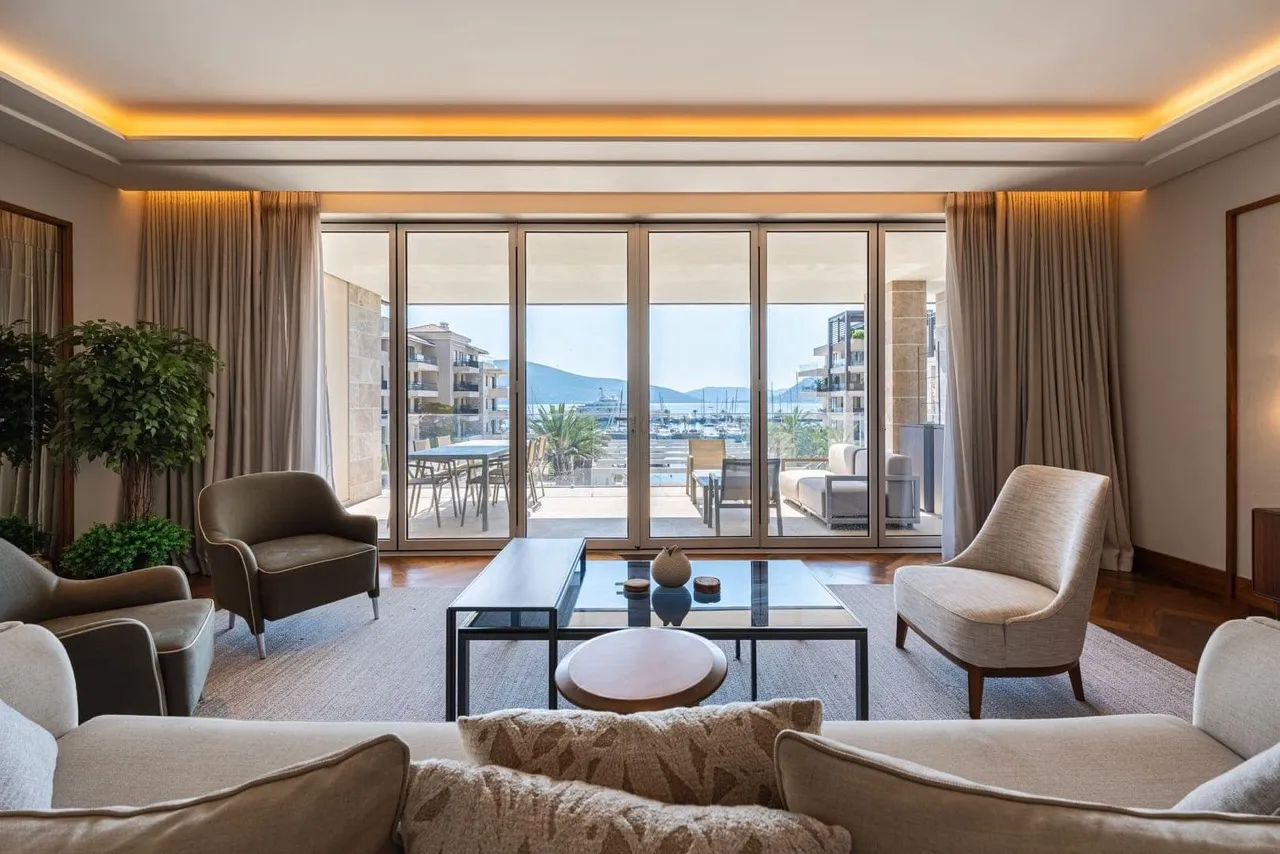 Bright living room with sofas, armchairs, and a glass table facing panoramic windows overlooking a marina.