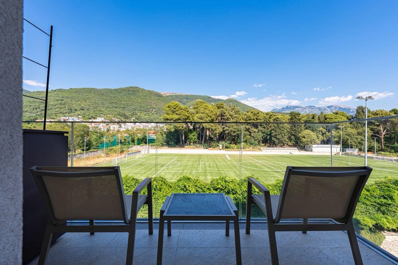 Apartment balcony in Porto Montenegro, Tivat, Montenegro with a view of the football field, mountains, and modern furniture.