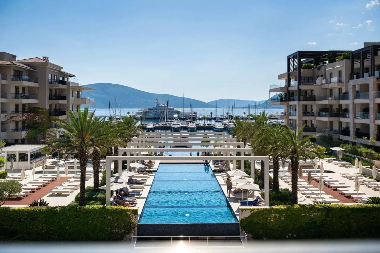 Large outdoor infinity pool with sunbeds and palm trees, overlooking a marina and sea.