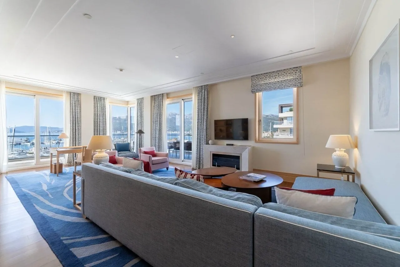 Bright living room featuring a large grey sofa, fireplace, and TV. A blue patterned rug covers the floor, with large windows overlooking a harbor.