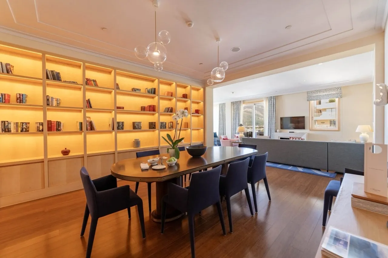 Living room featuring a wooden dining table, blue chairs, and a large illuminated bookshelf wall.