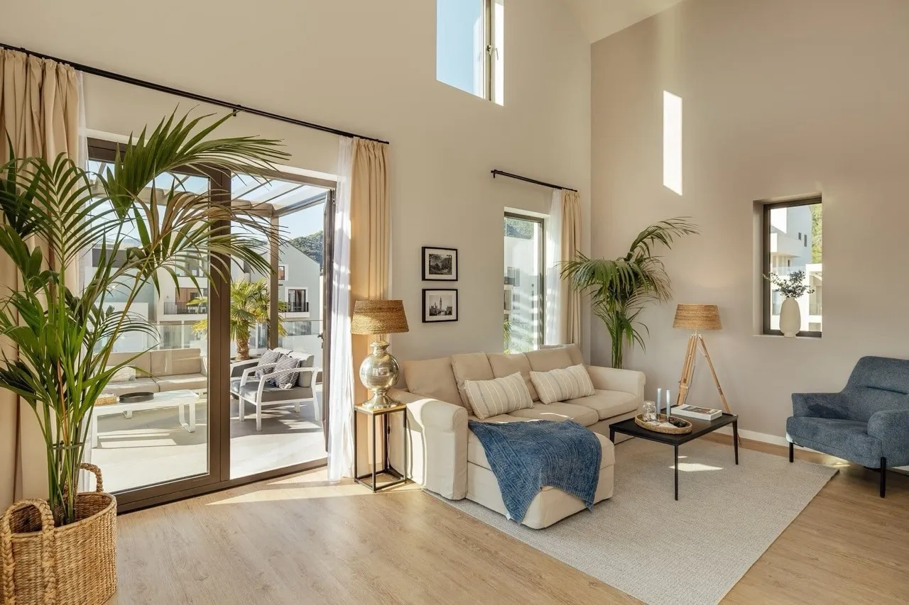 Bright living room with beige sofa, blue armchair, coffee table, and large plant near glass doors.