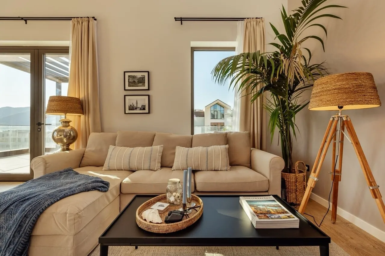 Bright living room featuring a beige sofa, black coffee table, tripod lamp, and tall indoor plant near the window.