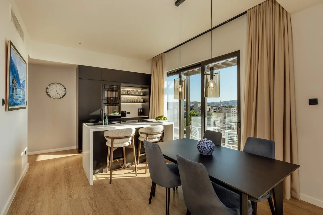 Modern kitchen with dark cabinets, white island, and dining area next to a large window.