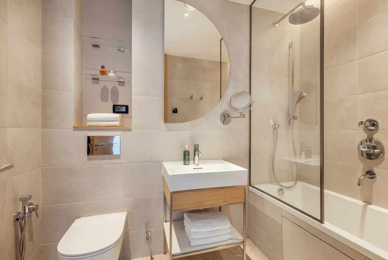 Modern bathroom with a vessel sink on a wooden vanity, glass shower enclosure, toilet, and round mirror on beige tiles.