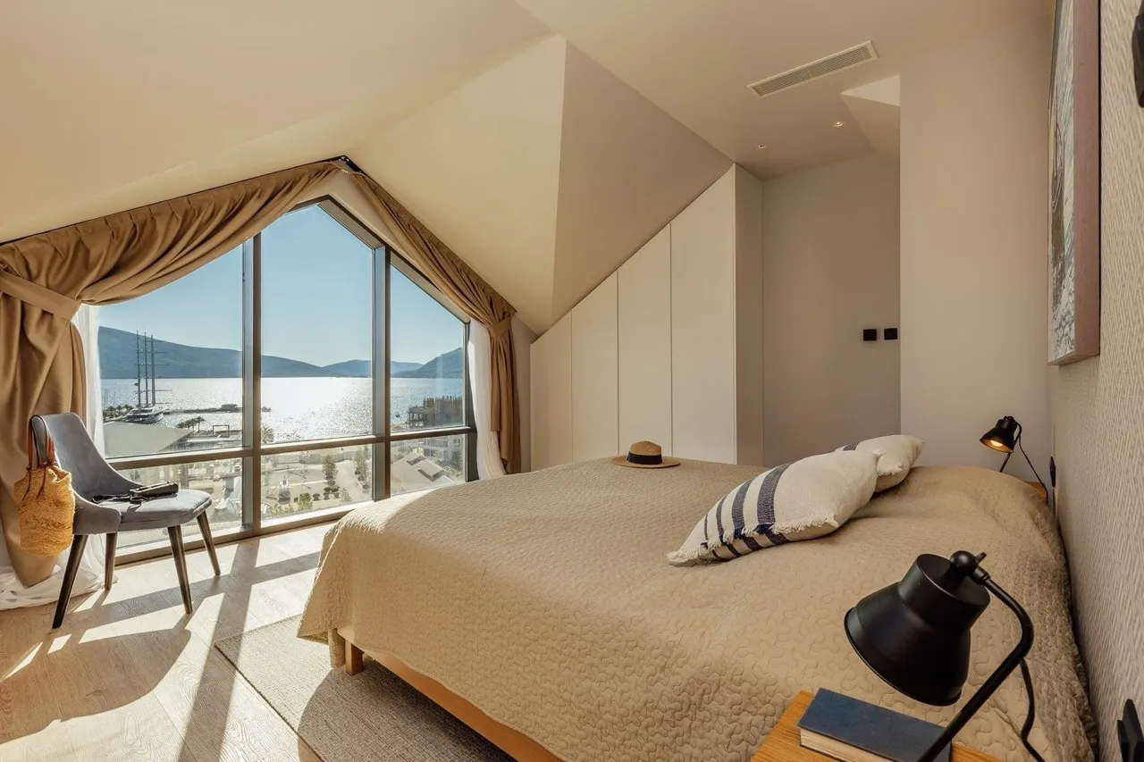 Bright bedroom with a large bed and panoramic window overlooking the sea and a marina.