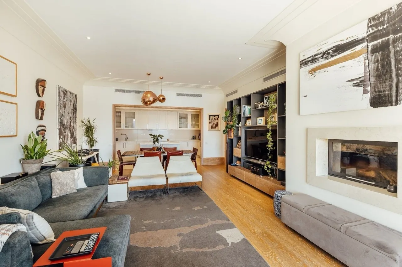 Bright living room with a grey sofa, fireplace, bookshelf, and an open kitchen visible in the background.