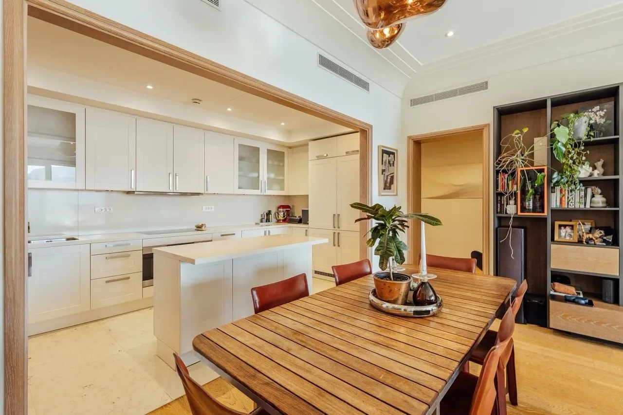 Modern kitchen with white cabinets and island, adjacent to a wooden dining table and bookshelf.