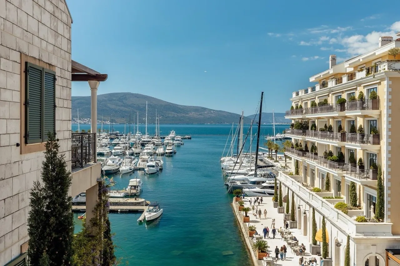 Panoramic view of a yacht marina, sea, and mountains from a balcony between buildings.
