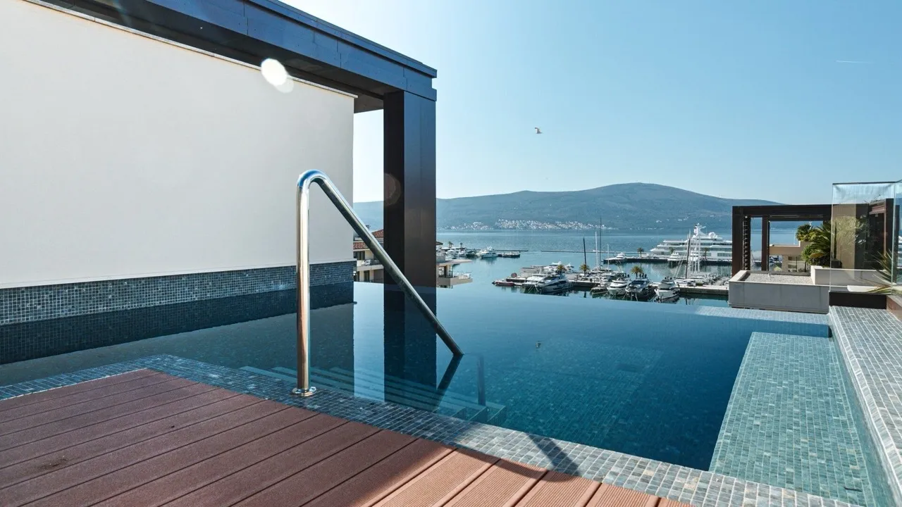 Infinity pool overlooking a marina and mountains. Features metal handrail, blue mosaic tiles, and wooden deck in foreground.