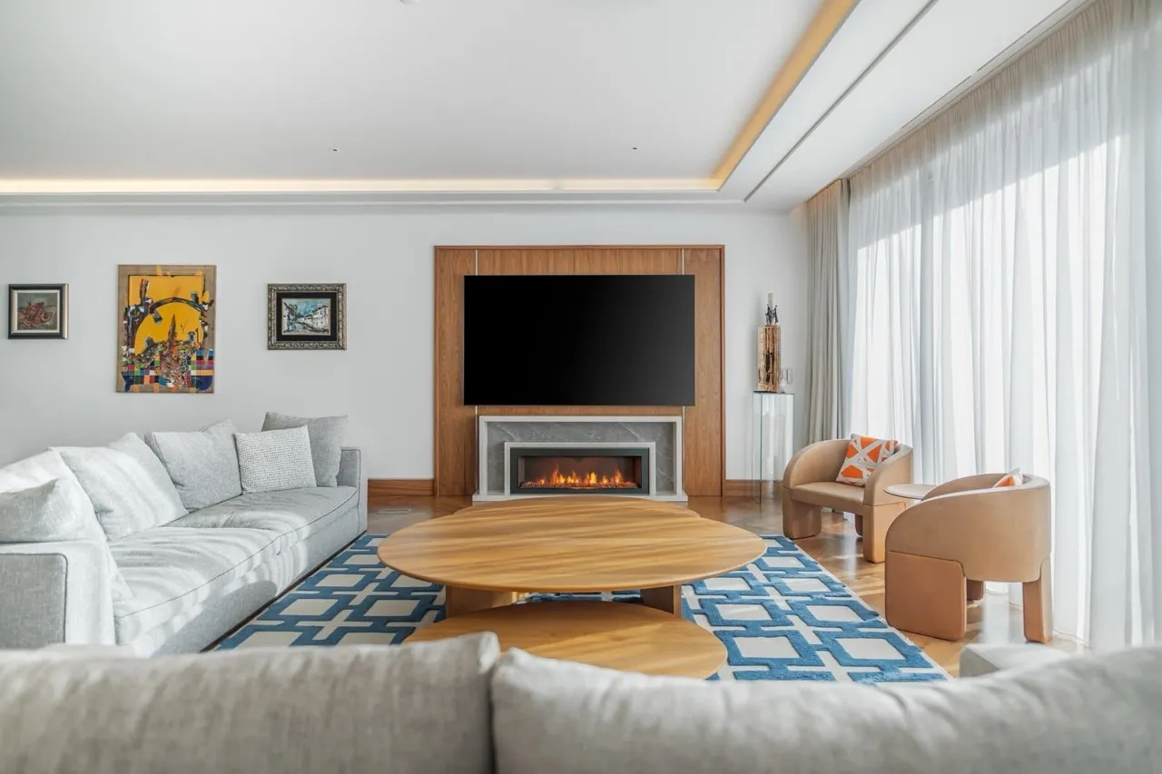 Bright living room with a grey sofa, wooden table, fireplace, and TV mounted on a wood panel wall.