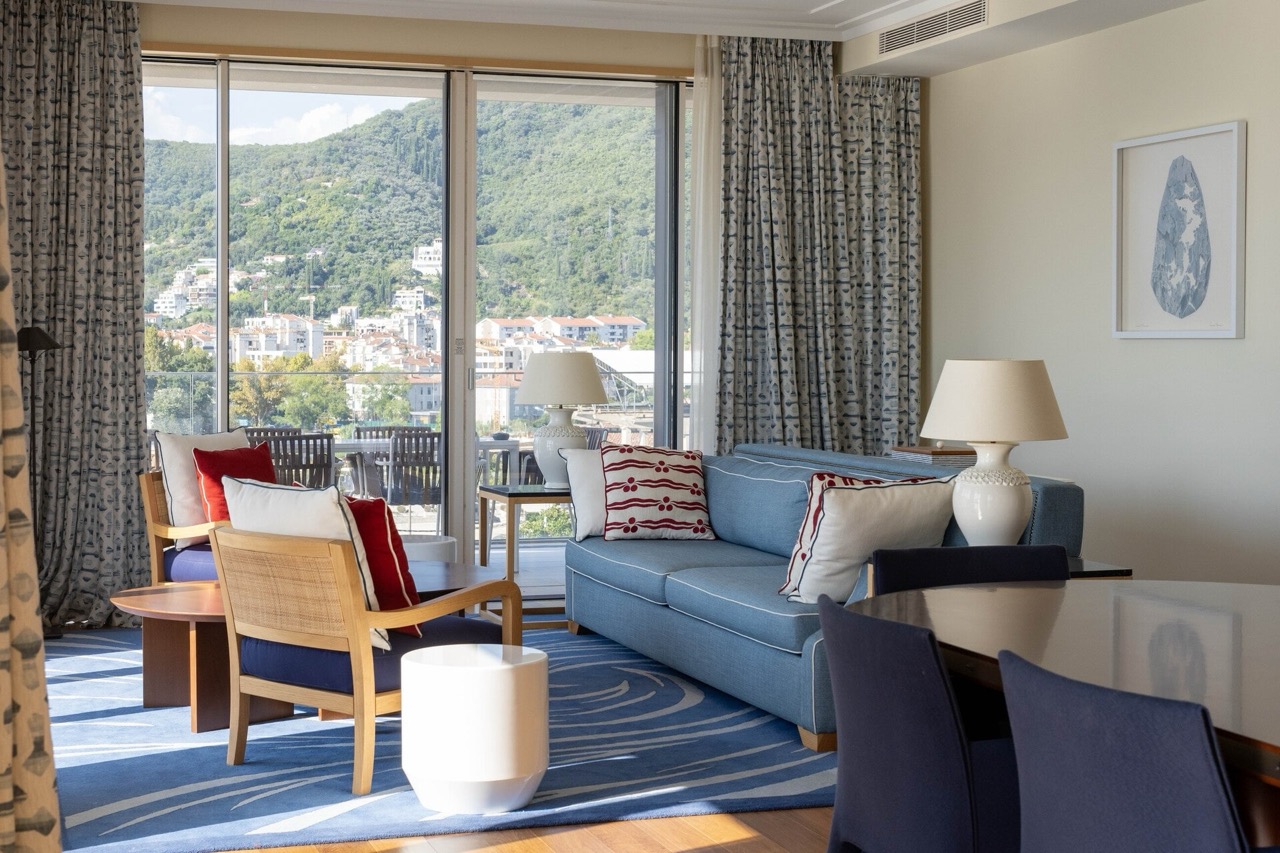 Living room in an apartment in Tivat, Montenegro: blue sofa, panoramic windows with mountain views, and a terrace with furniture.