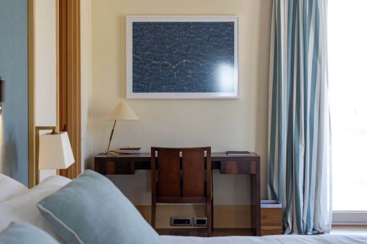 Bed with pillows, wooden desk, lamp, and framed art on the wall next to striped curtains.