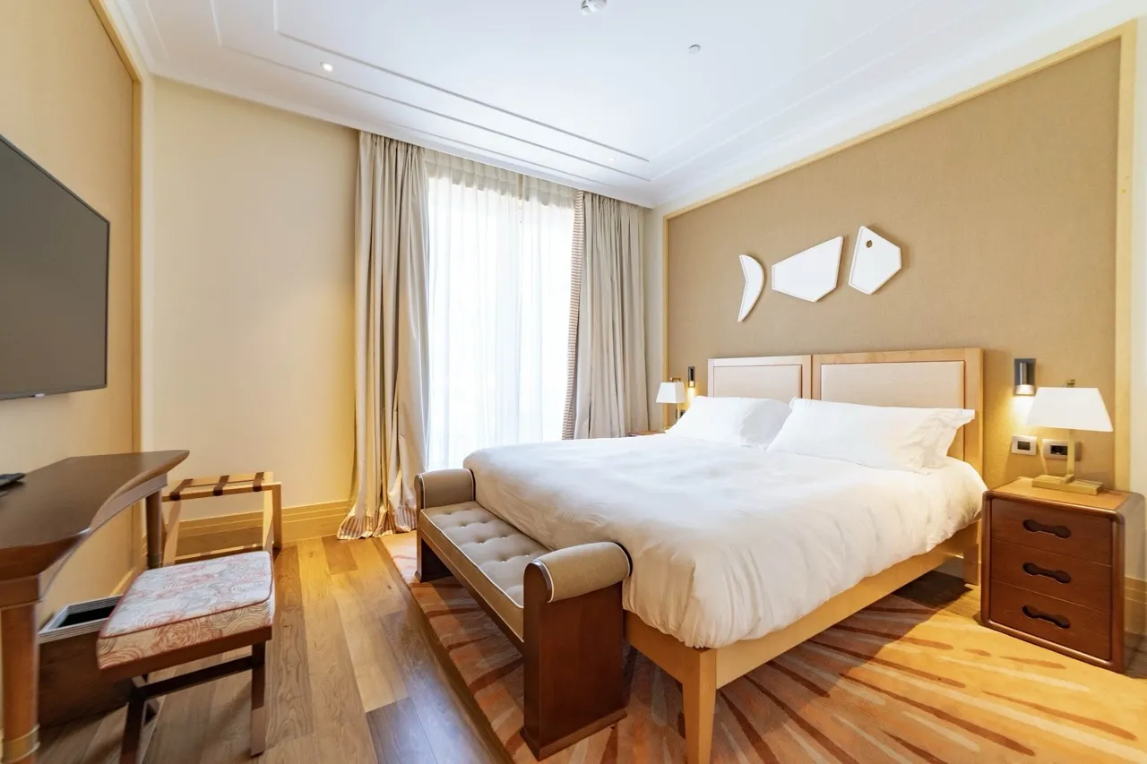 Bright bedroom with a large bed, wooden floor, nightstand with lamp, and wall-mounted TV.