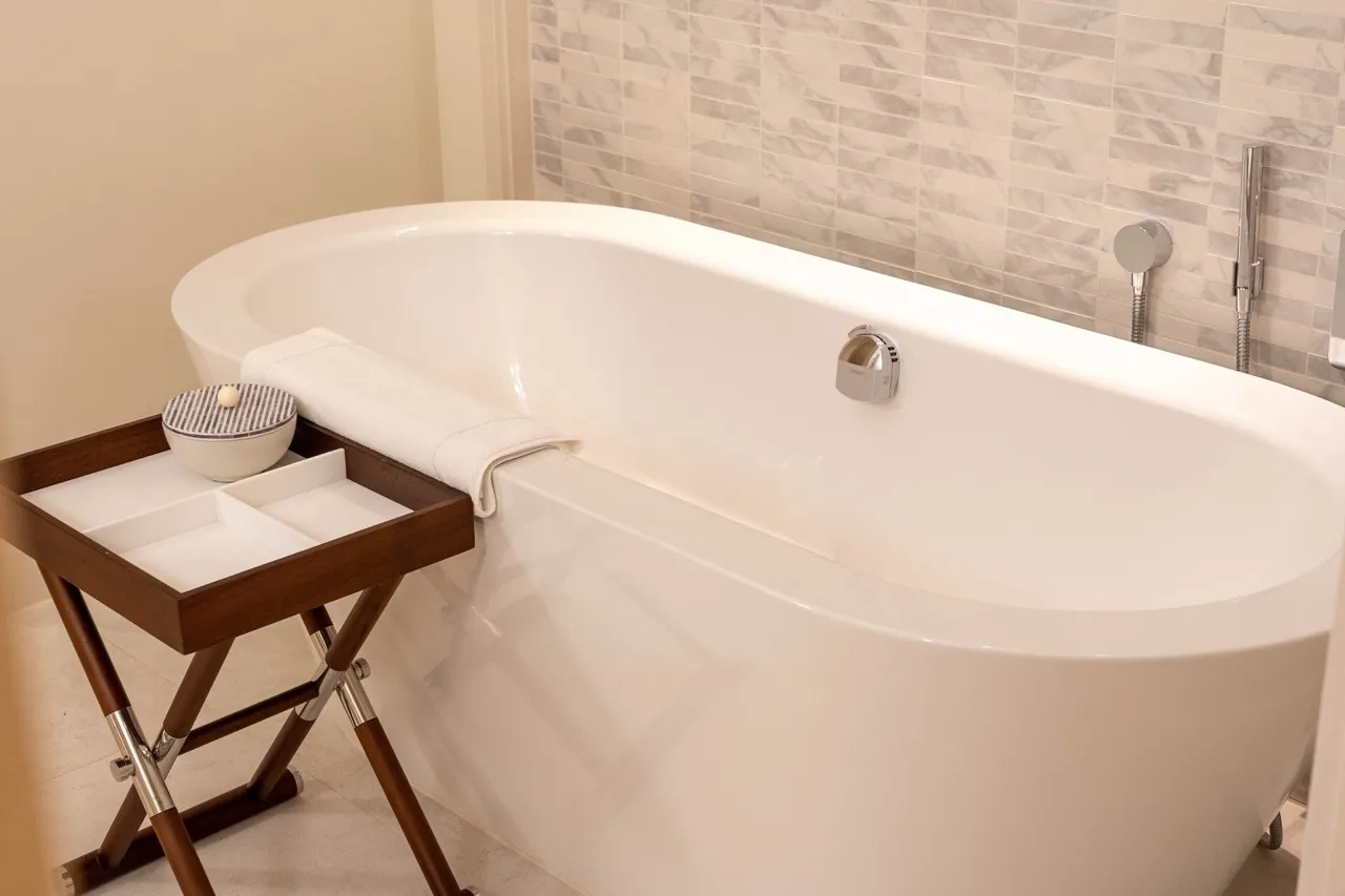 White freestanding bathtub, wooden caddy with towel and amenities, marble tile wall background.
