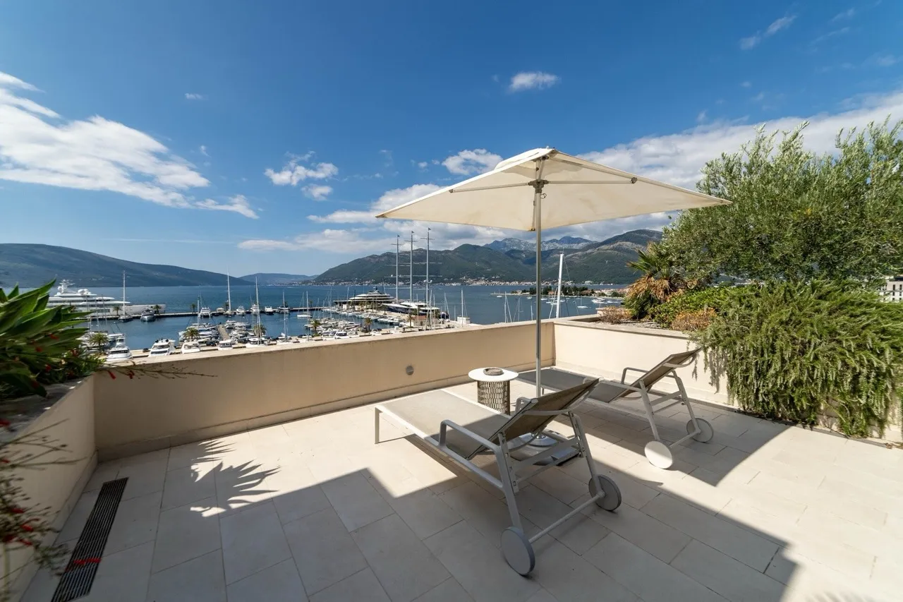 Terrace with sun loungers and umbrella overlooking a marina and mountains.