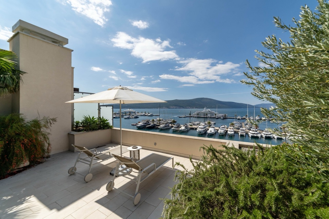 Apartment terrace in Tivat, Montenegro, with sun loungers, an umbrella, and a panoramic view of the Porto Montenegro yacht marina.