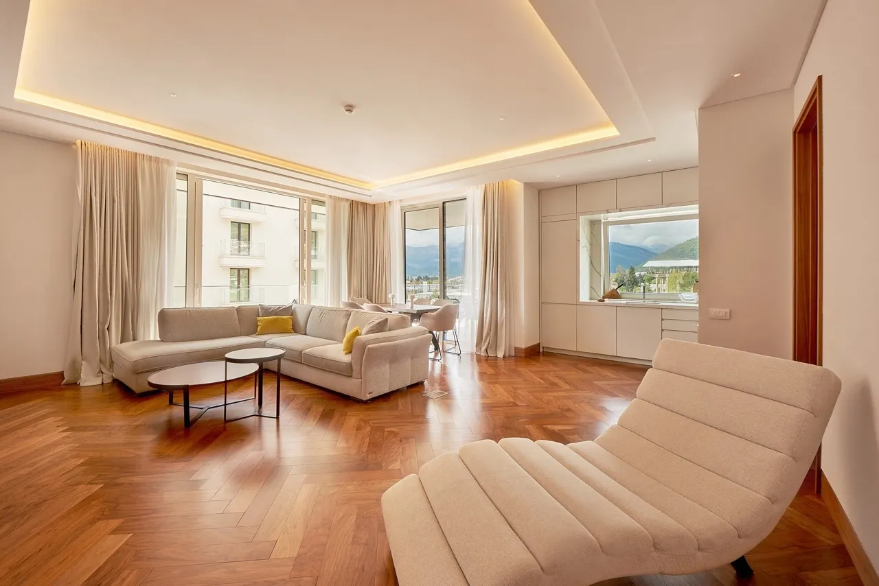 Bright living room with a beige sectional sofa, wooden parquet flooring, and an open kitchen visible in the background.