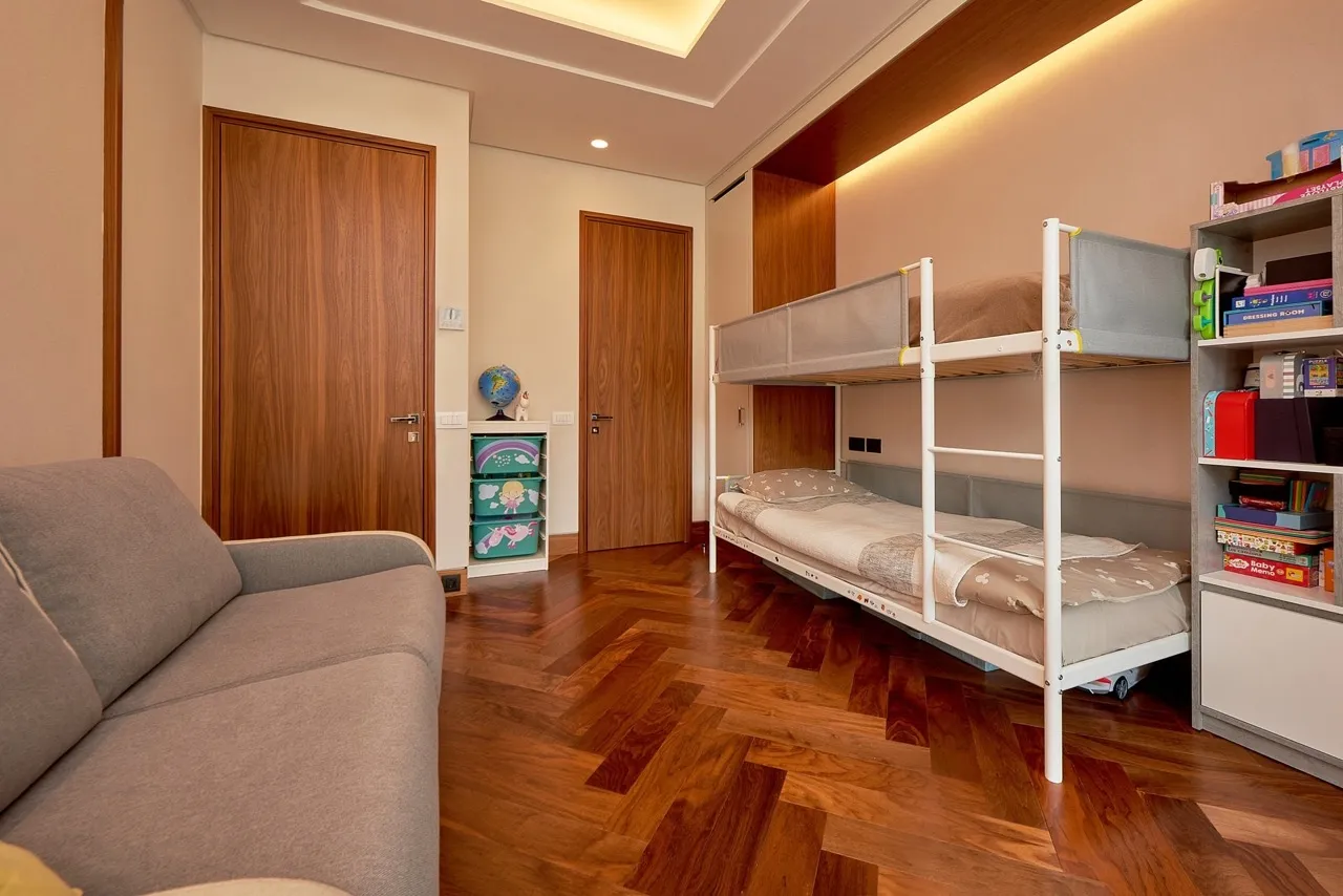 Children bedroom featuring a bunk bed, grey sofa, wooden parquet floor, and shelves with toys and books.