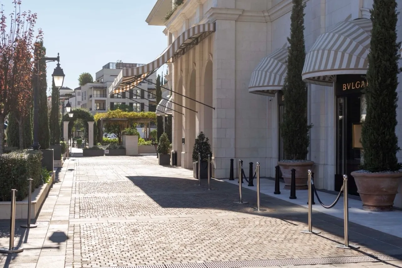 Paved outdoor walkway with striped awnings, arched architecture, and a Bulgari storefront. Features street lamps and cypress trees.
