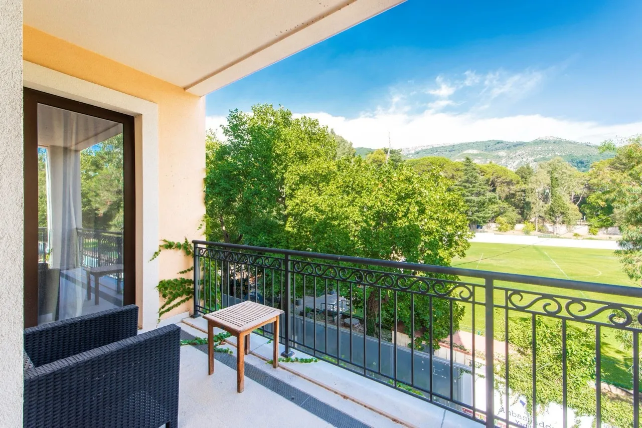 Balcony with black wicker chairs and a wooden table overlooking green trees and a sports field under a blue sky.