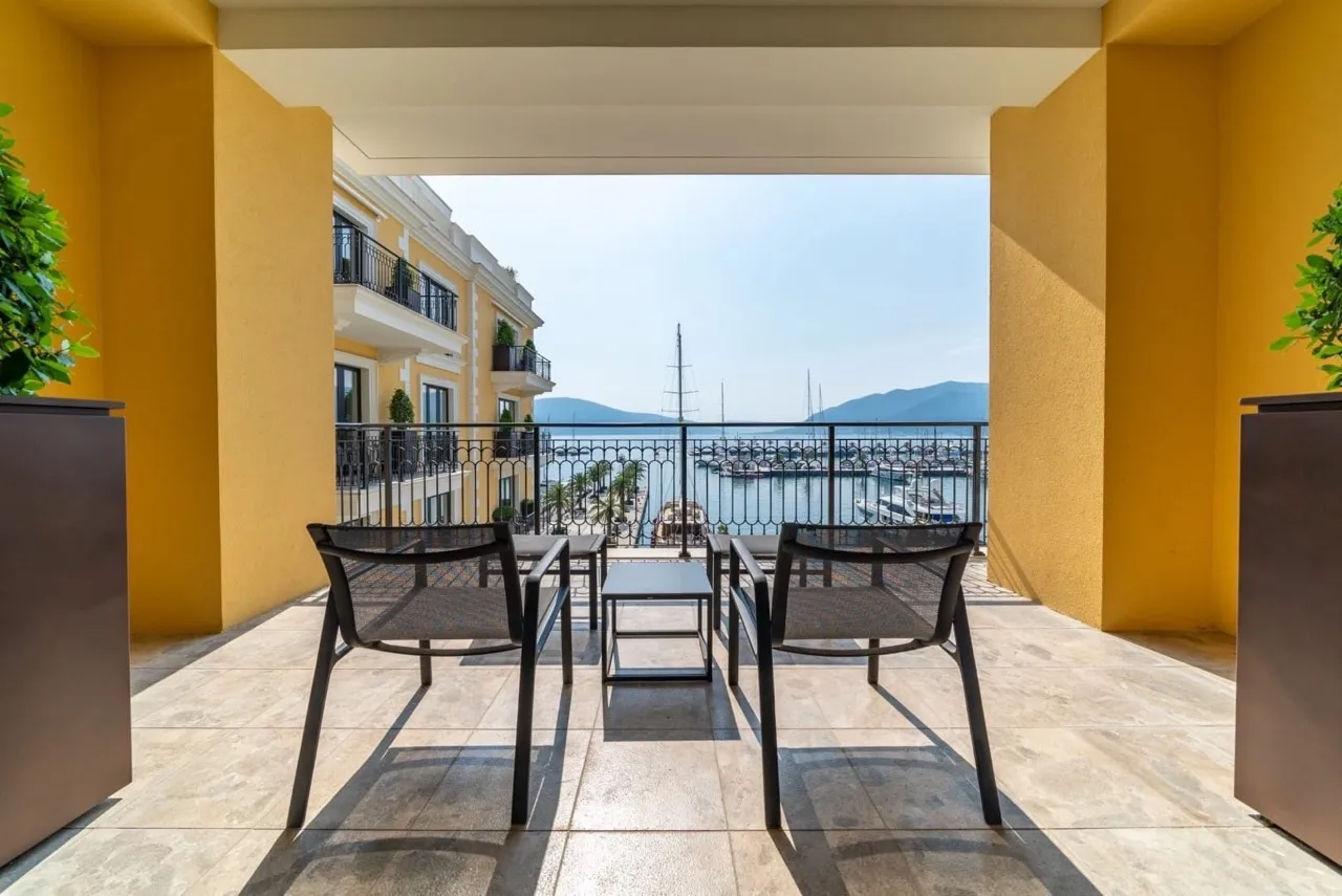 Balcony with black chairs overlooking a marina, featuring yellow walls, tiled floor, and iron railing.
