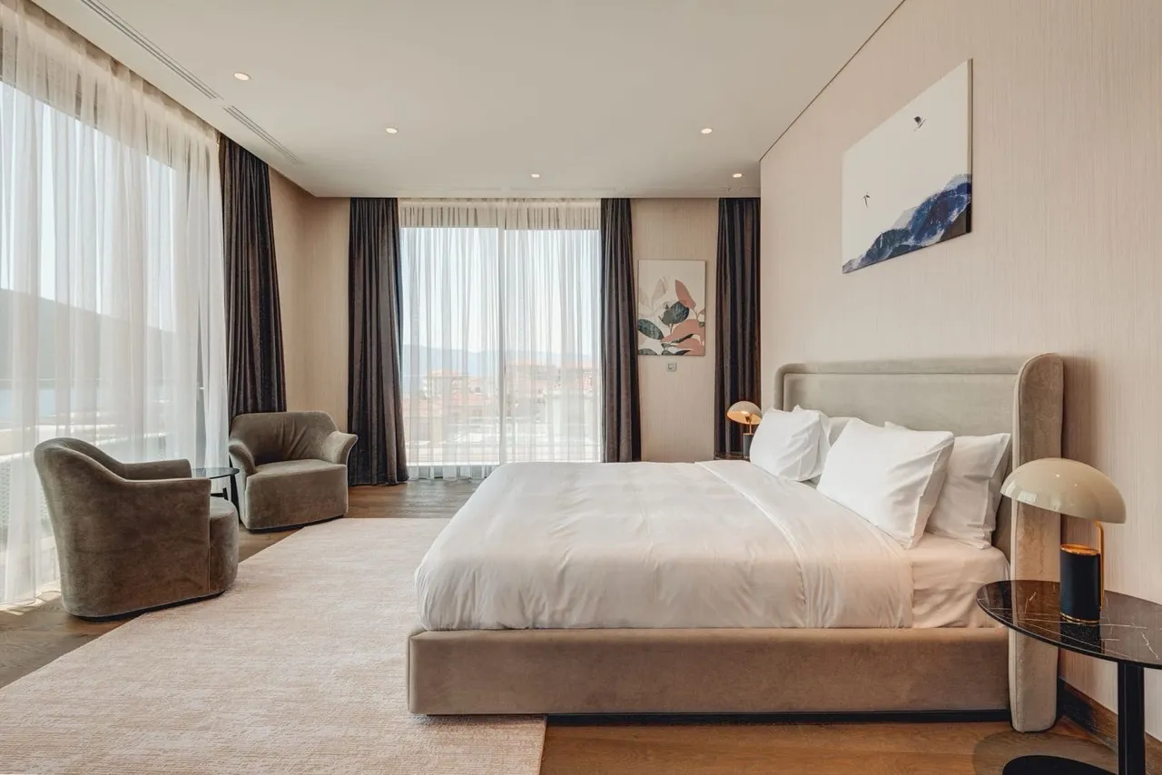 Modern bedroom featuring a large bed, velvet armchairs, and floor-to-ceiling windows.