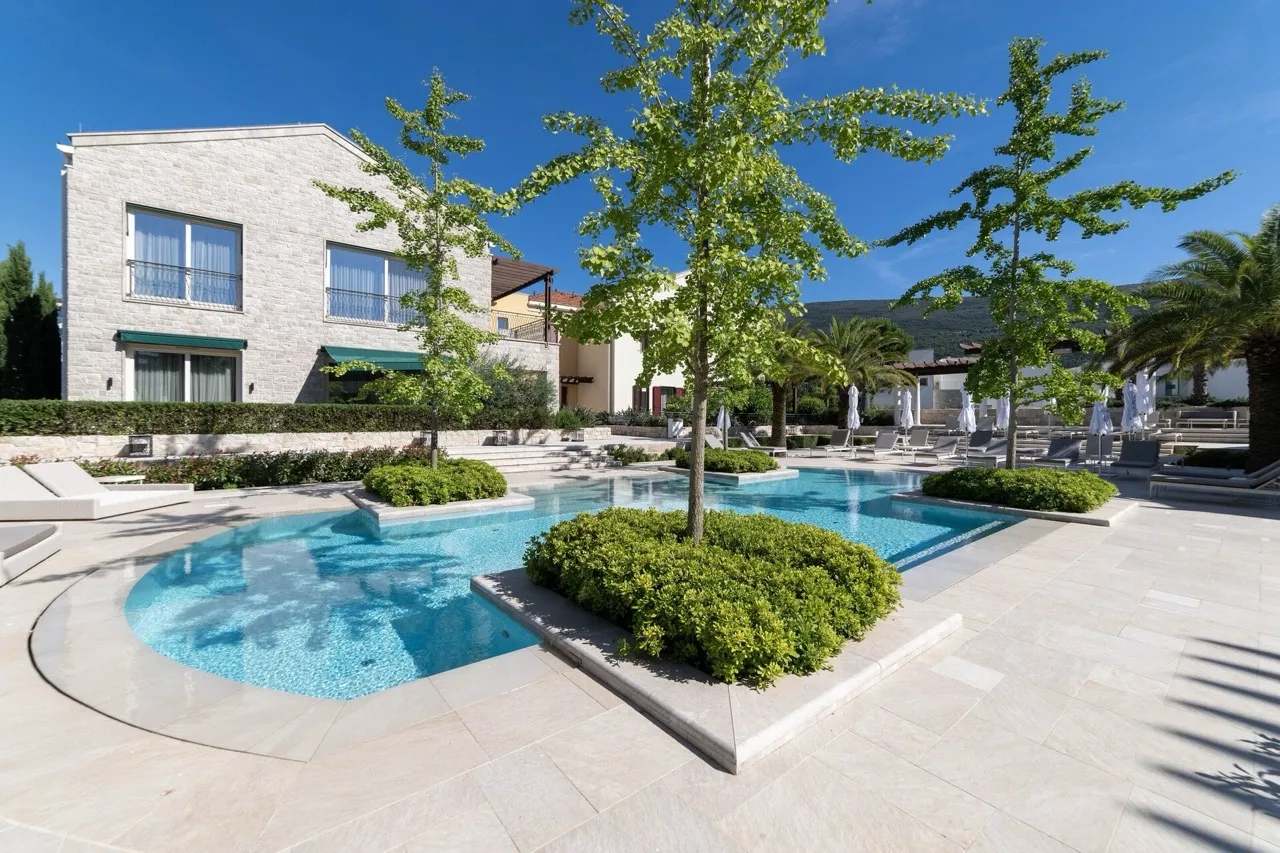 Outdoor swimming pool with blue water surrounded by stone decking, green bushes, and trees under a clear sky.