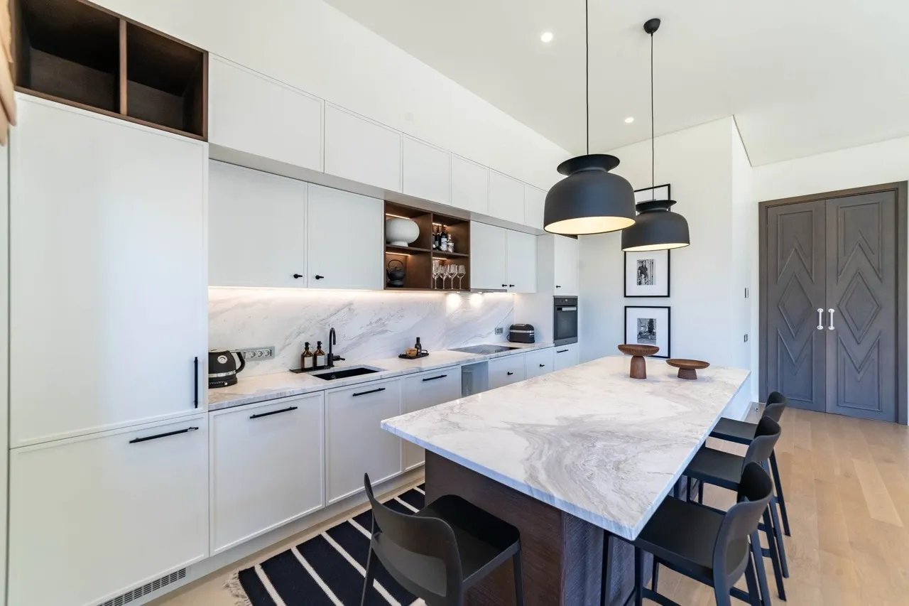 Modern kitchen with white cabinets, marble island, black bar stools, and pendant lights over the counter.