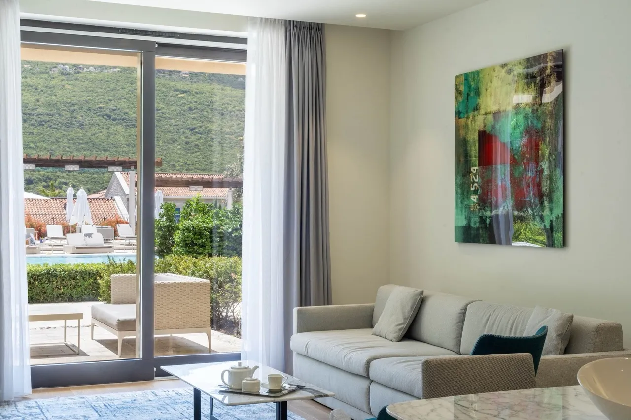 Bright room with a grey sofa, coffee table, and abstract art. A large sliding door opens to a terrace overlooking a pool and hills.