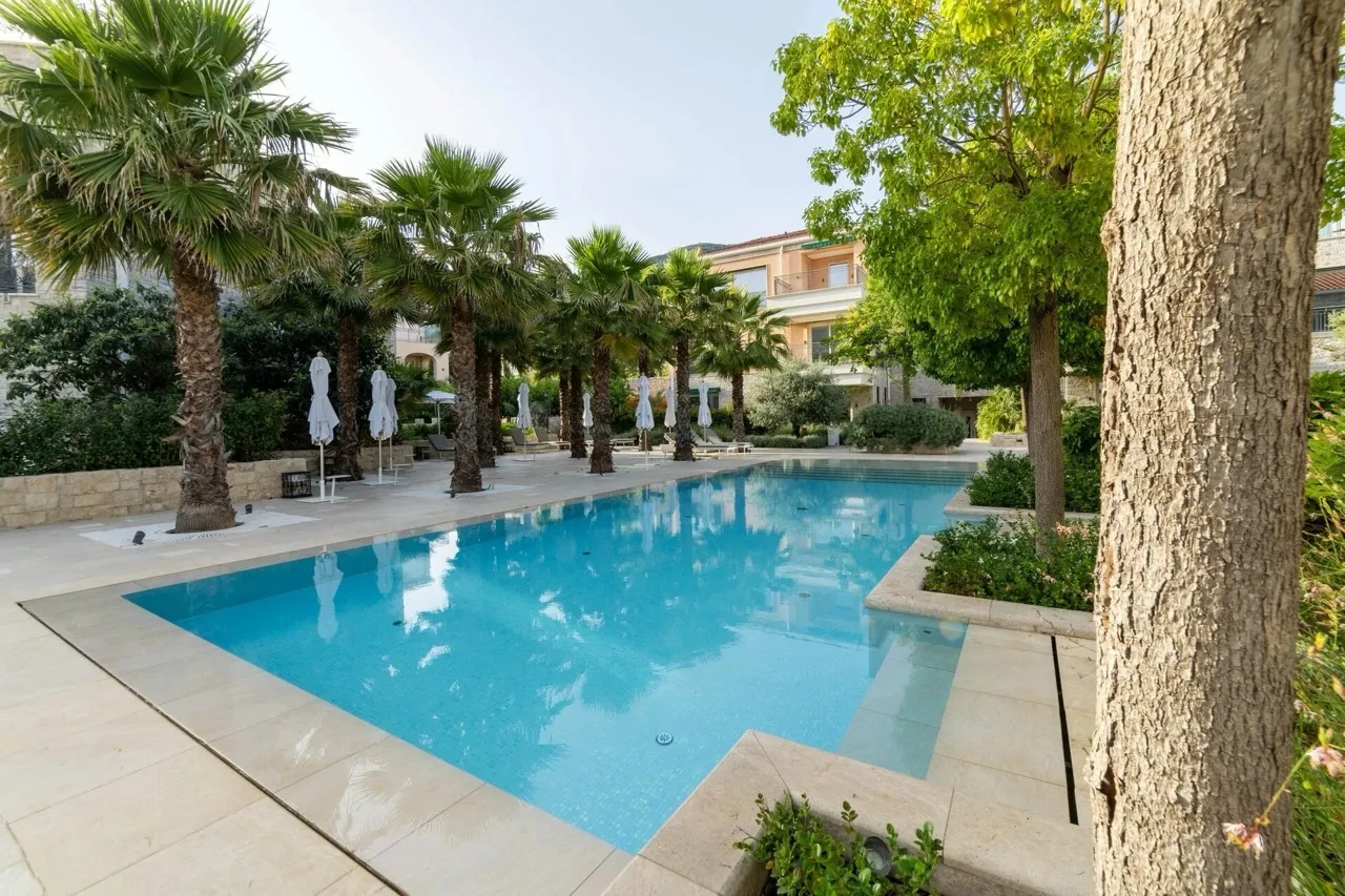 Outdoor swimming pool with blue water, surrounded by palm trees, closed umbrellas, and stone paving.