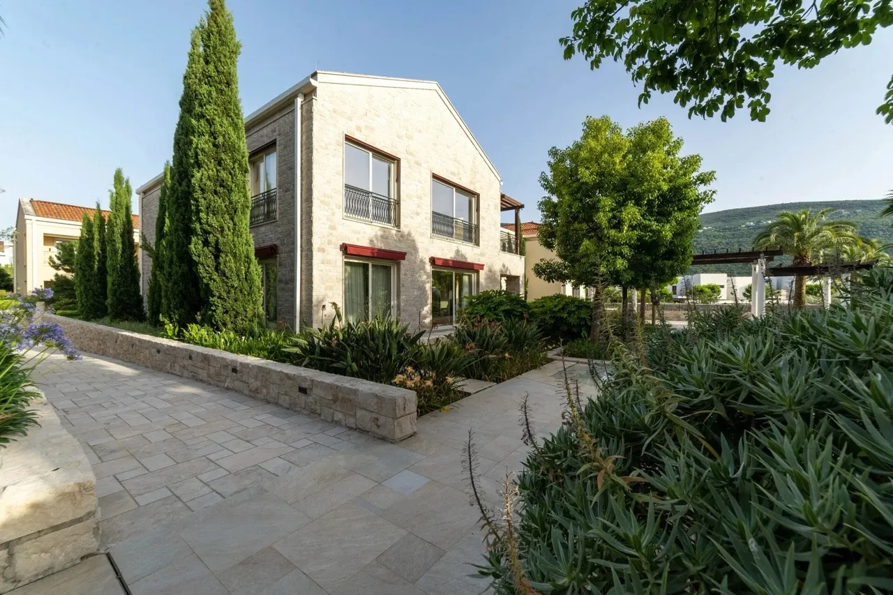Exterior of a stone house with a garden, paved walkway, tall cypress trees, and hills in the background.
