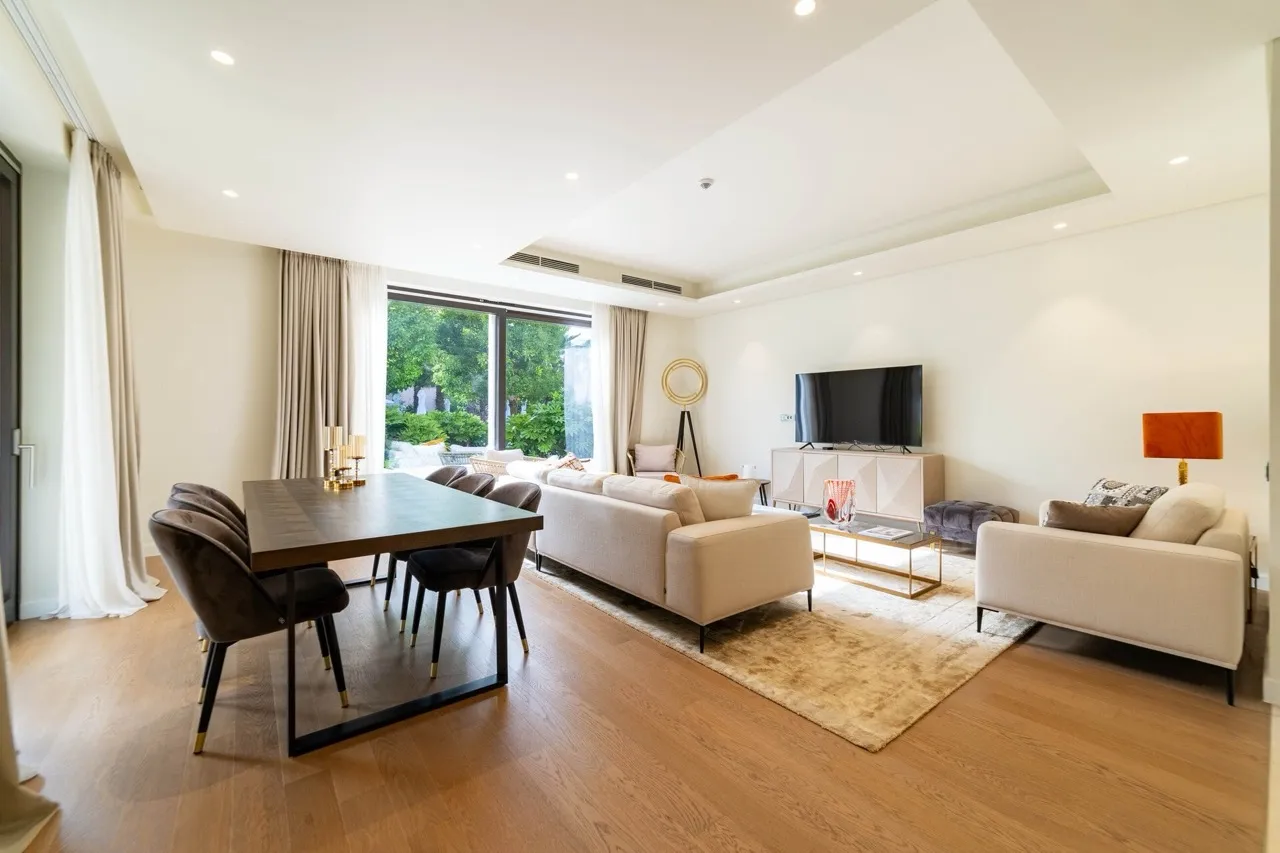 Bright living room with sofas, TV, and dining table on wooden flooring near large windows.
